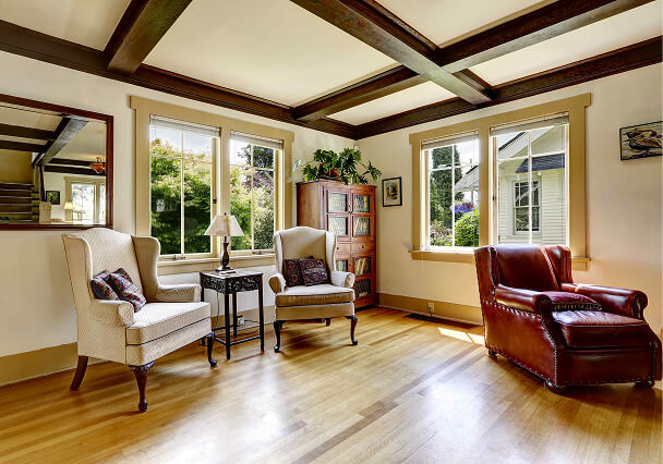 A cozy living room with two beige armchairs and a red leather armchair on a wooden floor, near sunlit windows and a bookcase, exuding warmth.