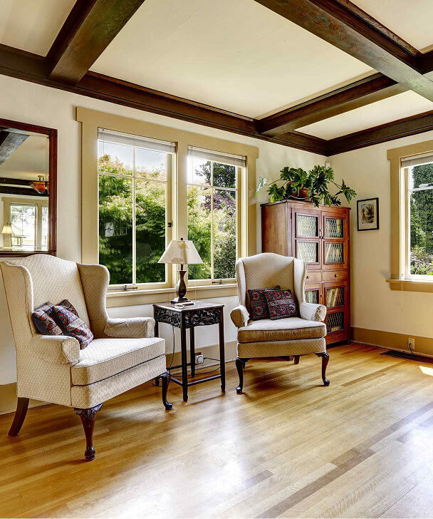 Elegant living room with two plush armchairs near a window. A vintage wooden cabinet, table, and plants create a cozy, welcoming atmosphere.