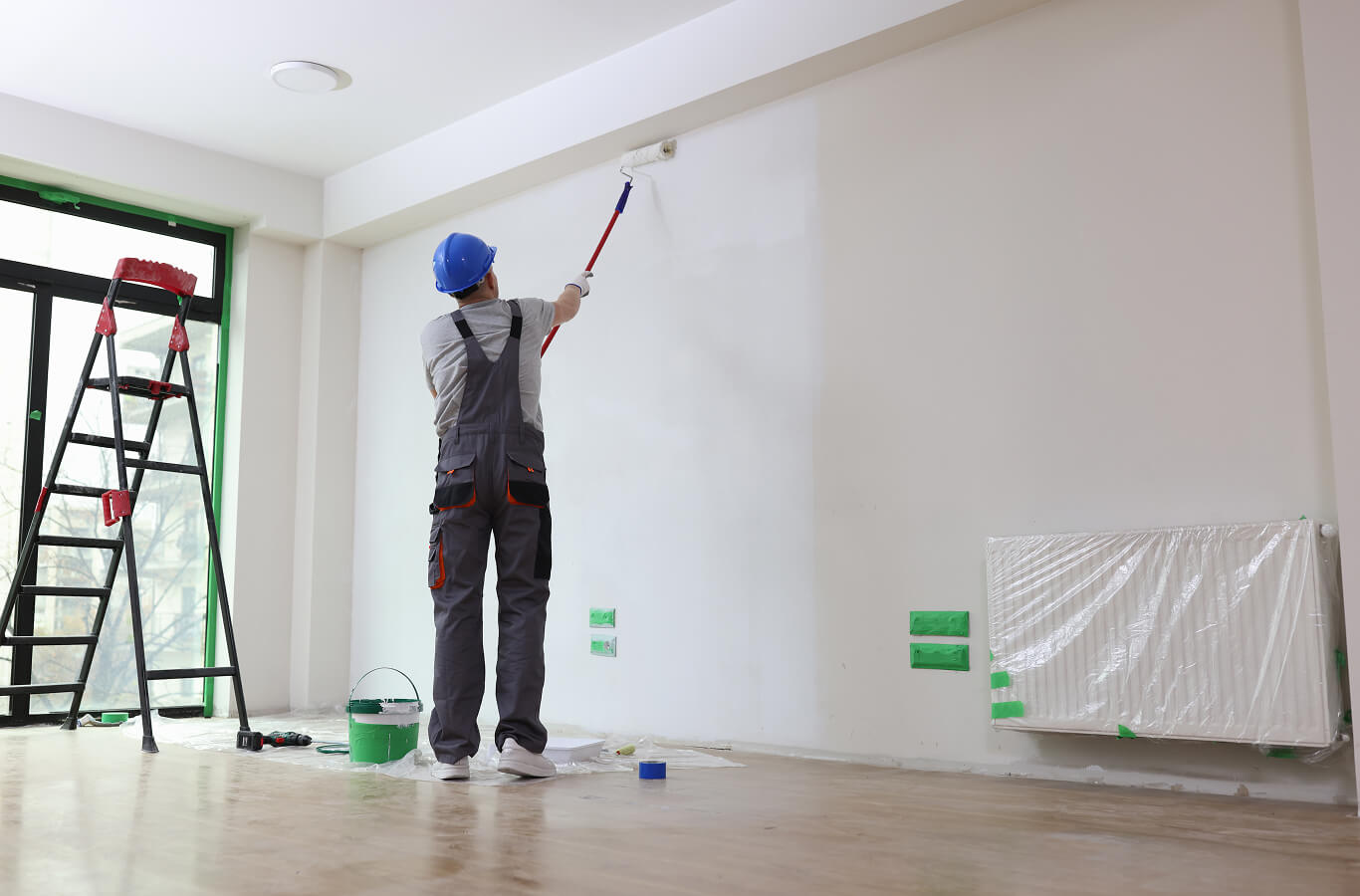 A worker in overalls and a hard hat paints a white wall with a roller. A ladder, paint bucket, and protective covers on the floor create a tidy, professional scene.