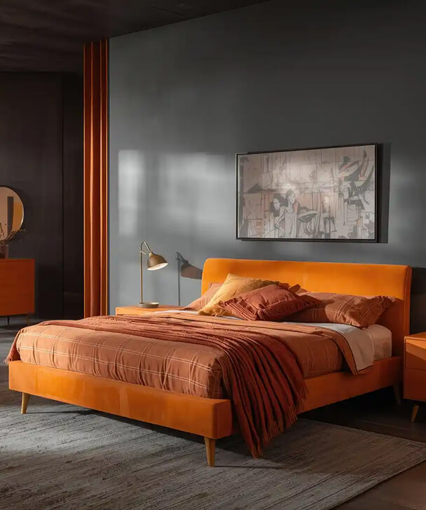 Cozy bedroom with an orange bed, layered with matching pillows and a textured throw. Soft lighting and abstract art create a warm, inviting ambiance.
