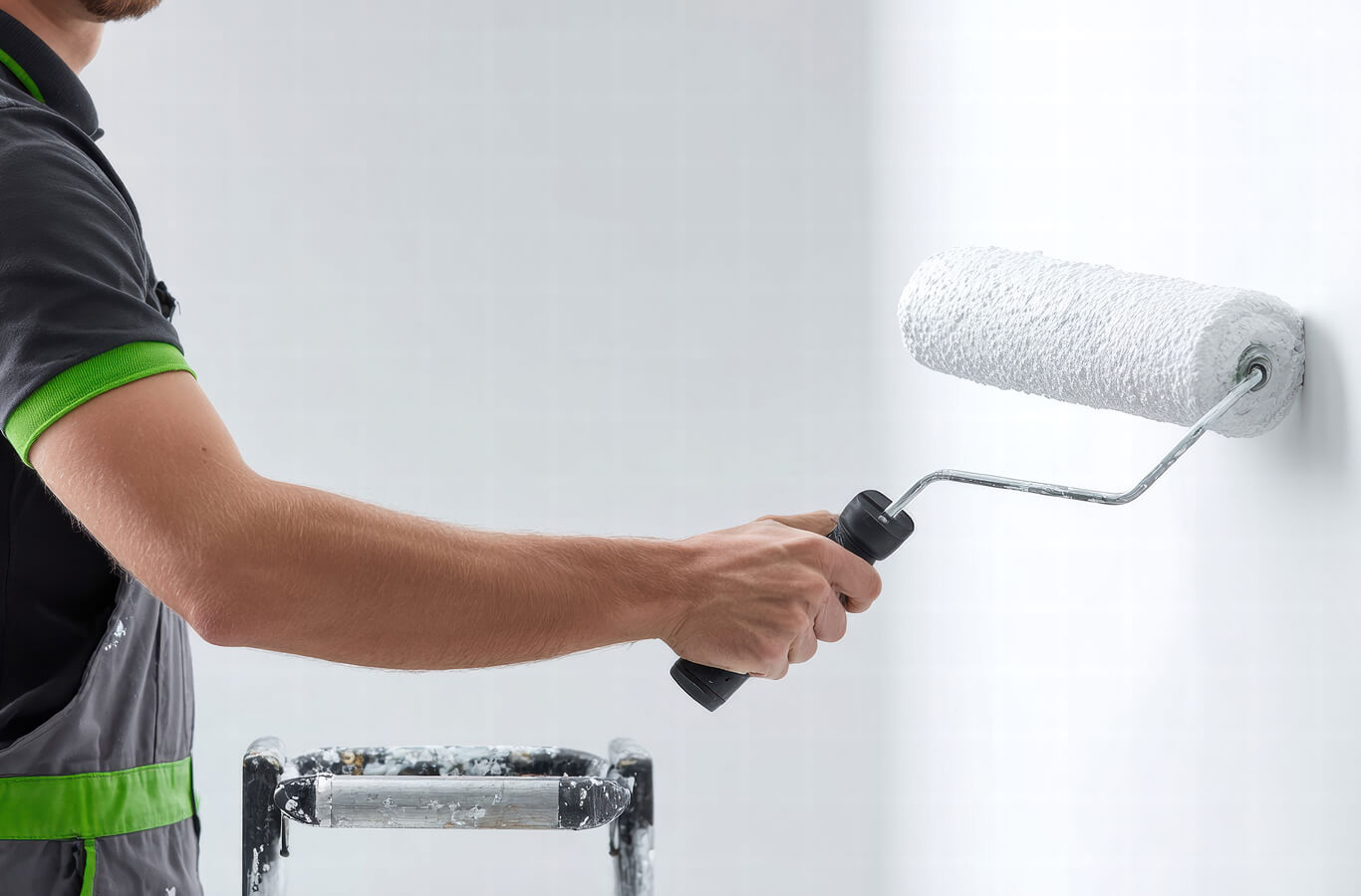 A person in a grey uniform with green trim uses a paint roller to apply white paint on a wall, next to a ladder with paint splatters.