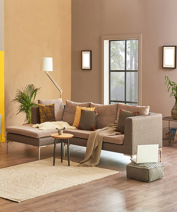 Modern living room with a beige L-shaped sofa adorned with assorted cushions and a throw. A floor lamp and potted plants add warmth. Two nested round tables sit on a textured rug. Large window lets in natural light, enhancing the cozy, minimalist ambiance.