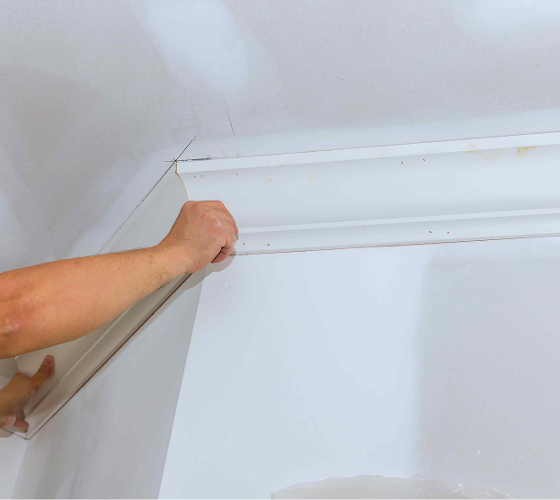 Hands are installing white crown molding on a ceiling corner. The scene conveys a sense of craftsmanship and home improvement in progress.