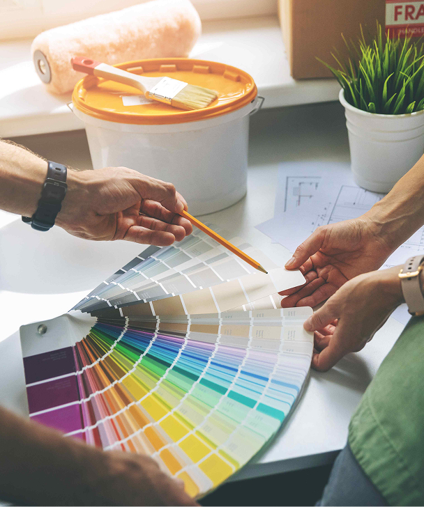 Two people examine a colorful paint swatch fan on a table, with a paint bucket, brush, roller, and plant nearby, suggesting a home renovation.