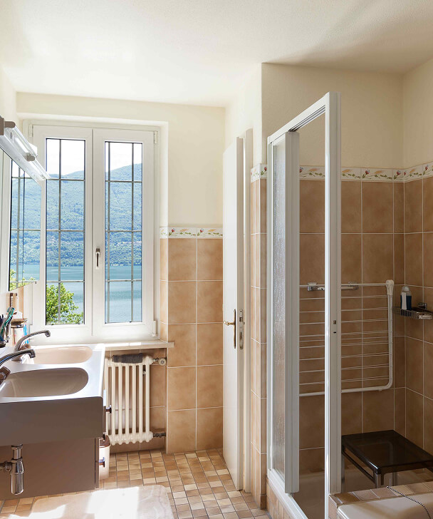 Bright bathroom with beige tiles, shower cabin, and a window overlooking a serene lake and mountains. Sunlight illuminates the room, creating a tranquil ambiance.