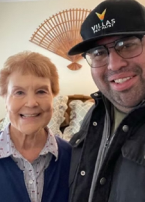 A smiling elderly woman in a blue cardigan and a man in glasses and a black cap stand together indoors, exuding warmth and happiness. Professional Painting Contractors.