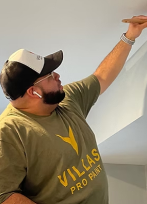 Man in green "Villas Pro Paint" shirt and cap, painting a wall with focus, conveying dedication. He wears AirPods and a watch, engaged in his work. Professional Painting Contractor.
