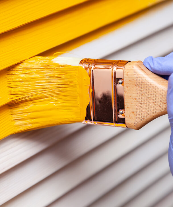 Close-up of a hand in a blue glove painting a white wooden surface bright yellow with a paintbrush. The scene conveys precision and transformation.